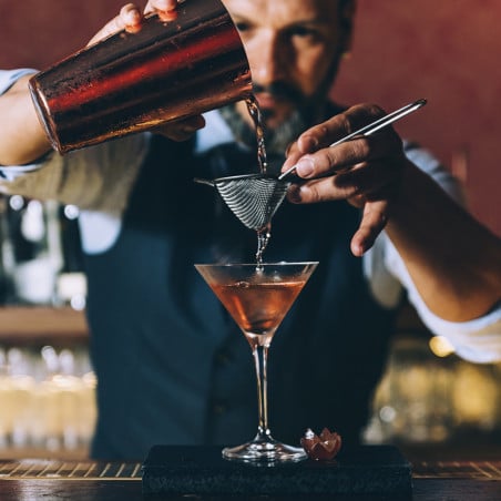 Bartender preparando cóctel en copa martini con licor ámbar vertido desde coctelera, vista frontal en barra